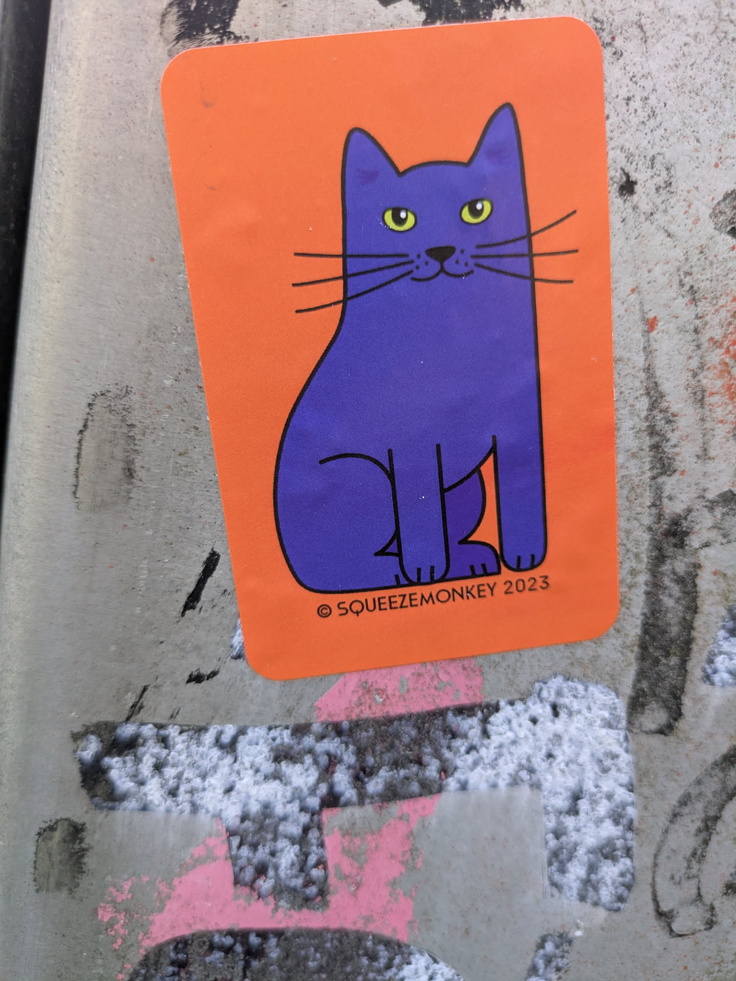 A rectangular sticker with rounded corners featuring a cartoon-style seated cat. The cat is a solid dark purple color with bright yellowish-green eyes, a small black nose, a subtle smile, and long black whiskers. The cat is outlined in black, and the background of the sticker is a vibrant orange. At the bottom right, small black text reads "© SQUEEZEMONKEY 2023". The sticker is adhered to a textured, grey urban surface, which has some faint pink and white markings.