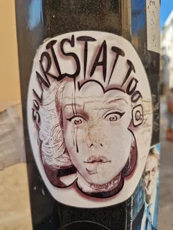Street sticker A sticker featuring a stylized illustration of a woman's face with a unique hairstyle and makeup. The style is reminiscent of graphic novels or vintage prints. The text 'Solaristattoo' is written in a graffiti-style font above the illustration, likely representing the artist's name or brand. There is also a small Instagram logo next to the text. The sticker appears to be applied to a dark-colored pole, potentially outdoors.