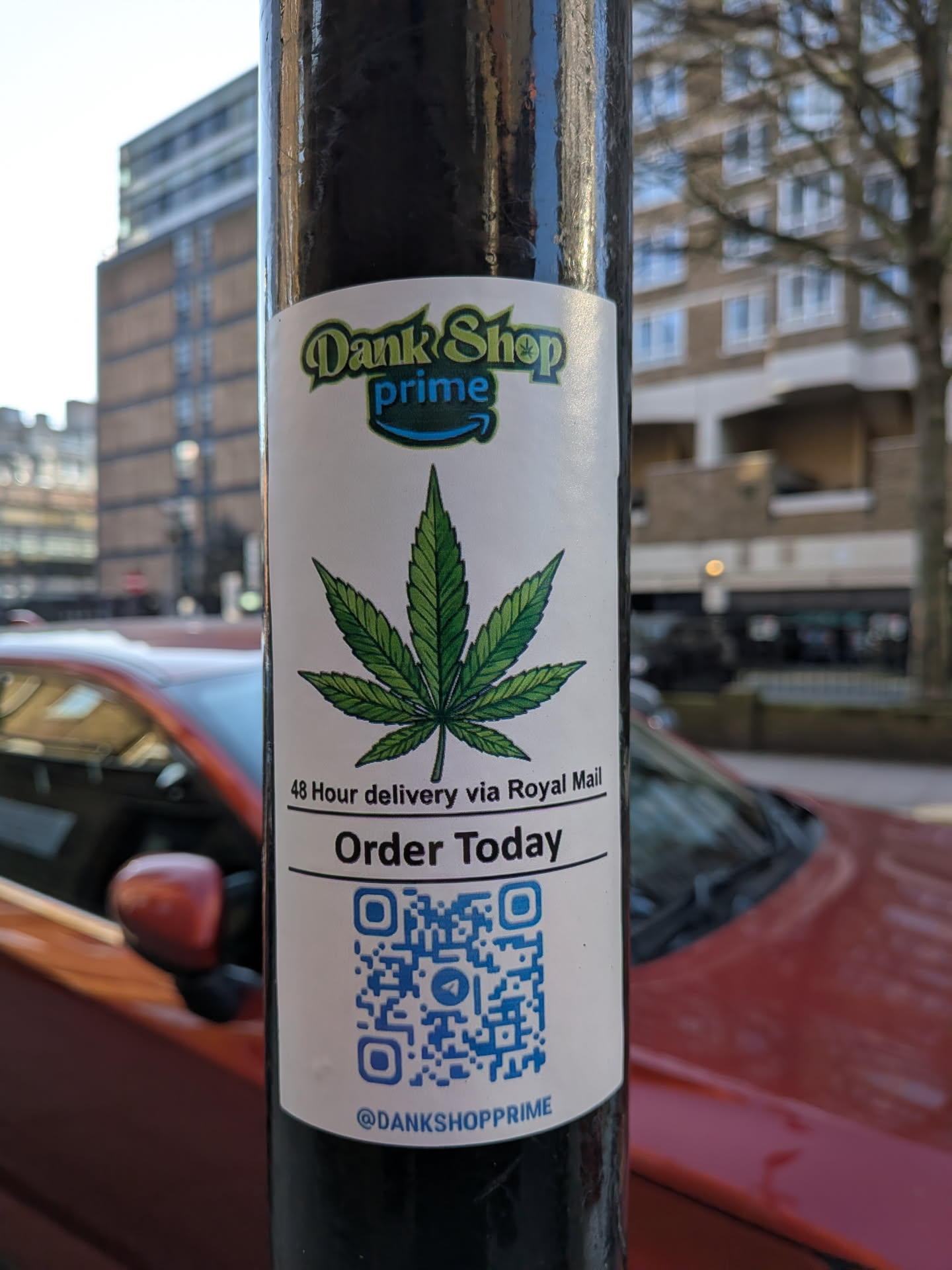 The sticker is a white, rectangular label with rounded corners, affixed to a black pole. At the top, it features a logo with 'Dank Shop' in a stylized green font, and 'prime' in blue, mimicking the Amazon Prime branding with an arrow swoosh underneath. Below the logo, a prominent, detailed illustration of a green cannabis leaf with seven serrated leaflets is centered. Underneath the leaf, a thin black line separates it from the text '48 Hour delivery via Royal Mail' in black. Another black line follows, above the phrase 'Order Today' in a bolder, larger black font. The lower section of the sticker contains a blue QR code, and at the very bottom, the social media handle '@DANKSHOPPRIME' is printed in small black text.