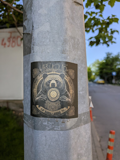A square sticker adhered to a metal pole features a detailed illustration of a bear holding a record in its hands. The artwork has an intricate, almost ornamental quality. The text "URSUDUB" is visible above the bear.