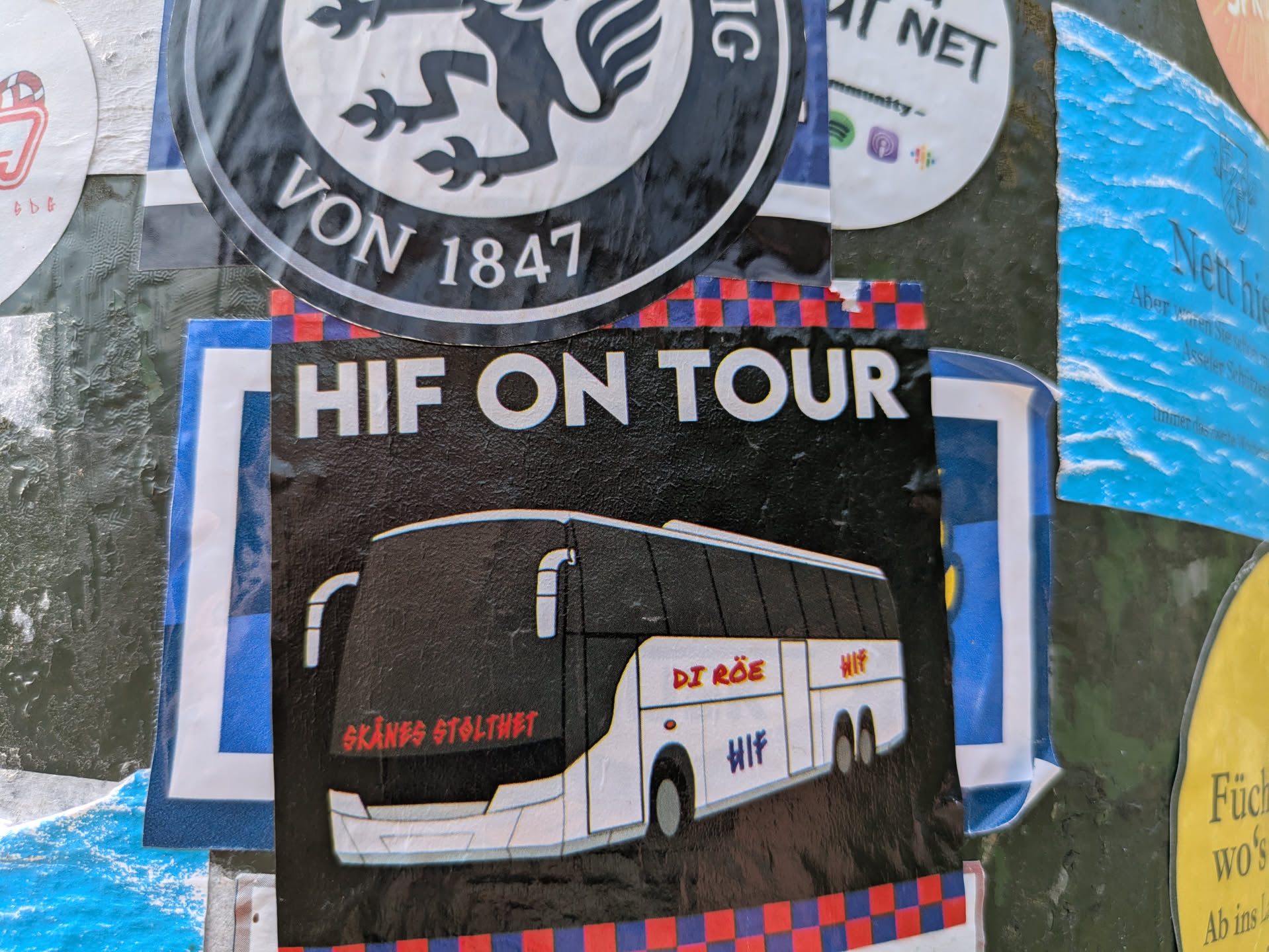 A rectangular sticker with a black background and a red and blue checkered pattern at the bottom. The top features the white text "HIF ON TOUR". Below this, there is an illustration of a white and black tour bus. On the front of the bus, in red text, it reads "SKÅNES STOLTHET" (Pride of Skåne). In the middle of the bus, also in red, it says "DI RÖE" (The Reds), and below that in a purple/blue shade, "HIF". At the rear of the bus, in red text, is "HIF". The sticker is dedicated to a football club and its supporters traveling.