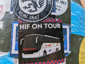 Street sticker A rectangular sticker with a black background and a red and blue checkered pattern at the bottom. The top features the white text "HIF ON TOUR". Below this, there is an illustration of a white and black tour bus. On the front of the bus, in red text, it reads "SKÅNES STOLTHET" (Pride of Skåne). In the middle of the bus, also in red, it says "DI RÖE" (The Reds), and below that in a purple/blue shade, "HIF". At the rear of the bus, in red text, is "HIF". The sticker is dedicated to a football club and its supporters traveling.