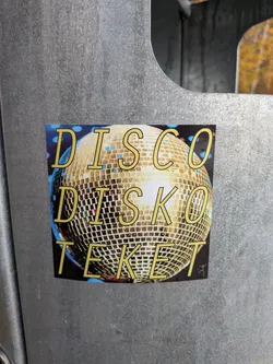 Street sticker The sticker features a prominent disco ball in the center, rendered with a golden, reflective surface composed of numerous small squares. The background is dark blue with subtle blue and yellow speckles, suggesting a vibrant, energetic atmosphere. Overlaying the disco ball are three lines of text in a bold, yellow, sans-serif font with a black outline. The top line reads 'DISCO', the second line reads 'DISKO', and the bottom line reads 'TEKET'. In the bottom right corner of the sticker, there is a small, stylized silhouette of a stick figure dancing.