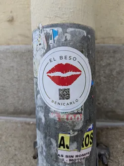 Street sticker This round sticker features a graphic of red lips in the center, with the text "EL BESO" above and "BENICARLÓ" below. A QR code is also printed below the lips. The sticker is adhered to a weathered, gray pole among other peeling stickers.