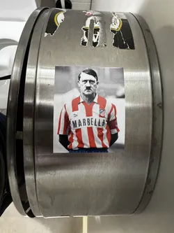 Street sticker The sticker displays a black and white image of Adolf Hitler, recognizable by his distinctive mustache and hairstyle, from the chest up. His torso is colorized, showing him wearing a red and white striped football jersey with a collar. The word 'MARBELLA' is prominently displayed in dark capital letters across the chest. A small Puma logo is visible on the right side of the chest, and a club crest, resembling that of Atlético Madrid, is on the left. A red armband with an indistinct dark symbol is visible on his right sleeve. The background of the figure is a blurred light grey, and the sticker itself is rectangular with a thin white border.