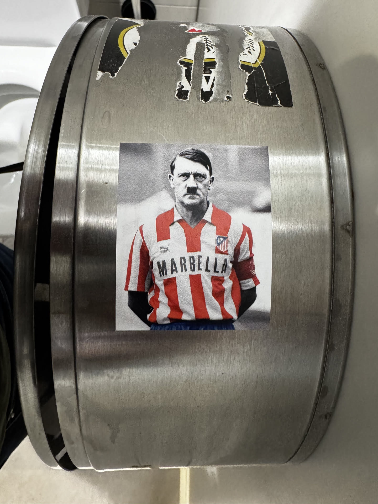 The sticker displays a black and white image of Adolf Hitler, recognizable by his distinctive mustache and hairstyle, from the chest up. His torso is colorized, showing him wearing a red and white striped football jersey with a collar. The word 'MARBELLA' is prominently displayed in dark capital letters across the chest. A small Puma logo is visible on the right side of the chest, and a club crest, resembling that of Atlético Madrid, is on the left. A red armband with an indistinct dark symbol is visible on his right sleeve. The background of the figure is a blurred light grey, and the sticker itself is rectangular with a thin white border.
