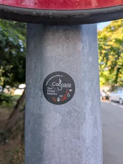 Street sticker A round black sticker on a metal post. It displays the 'Cascara Coffee Roastery' logo in white, the slogan 'Your Private Roastery', and the website 'www.cascaracoffee.ro'. Small orange and white coffee icons are arranged at the bottom.