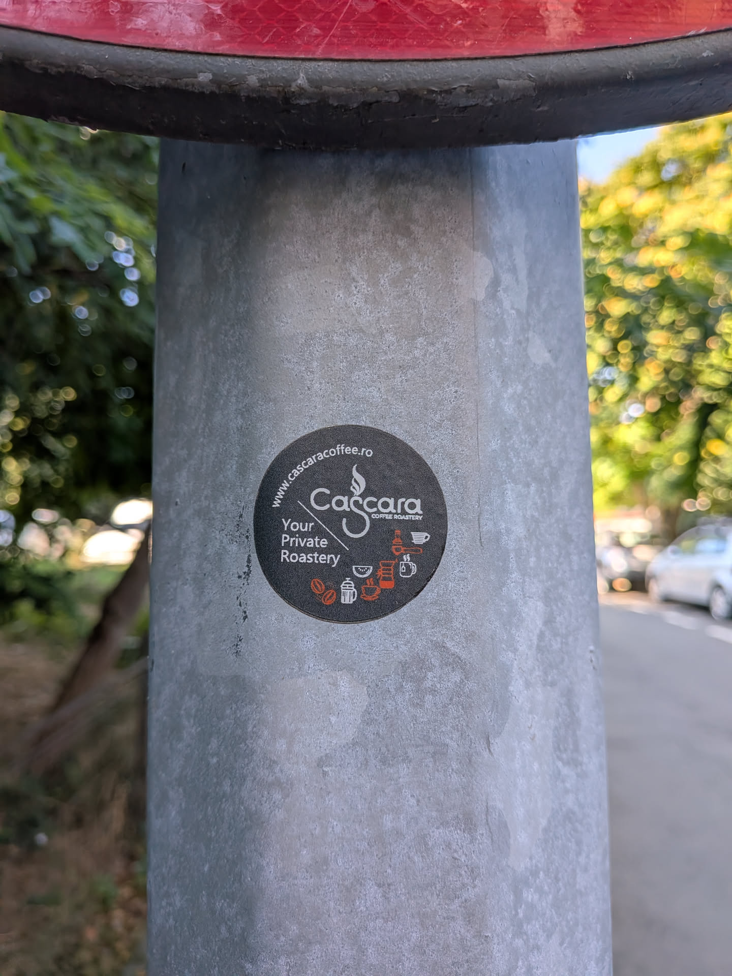 A round black sticker on a metal post. It displays the 'Cascara Coffee Roastery' logo in white, the slogan 'Your Private Roastery', and the website 'www.cascaracoffee.ro'. Small orange and white coffee icons are arranged at the bottom.