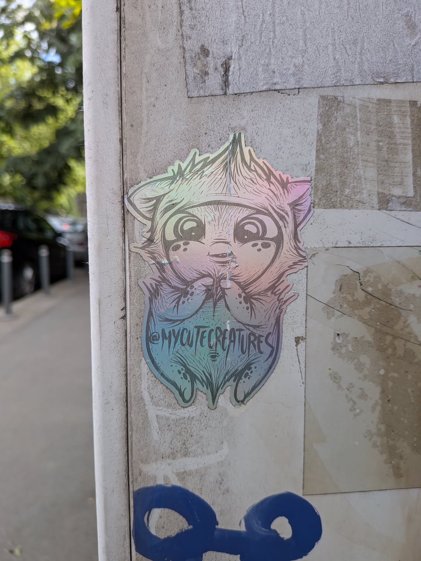A die-cut iridescent sticker depicting a cute, furry creature with large eyes and paws held together. Features black line art over a rainbow metallic background, with the handle @MYCUTECREATURES printed on the bottom.