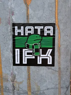 Street sticker The sticker is predominantly black with white text and a green and black striped scarf around a cartoonish green character. The text 'HATA' is at the top in large white block letters, and 'IFK' is at the bottom, also in large white block letters. Between the text, there is a horizontal green band. The character is green with small black dots for eyes and a simple mouth, wearing a scarf with alternating black and green stripes. The sticker appears to be slightly creased, indicating it is a physical sticker applied to a surface.