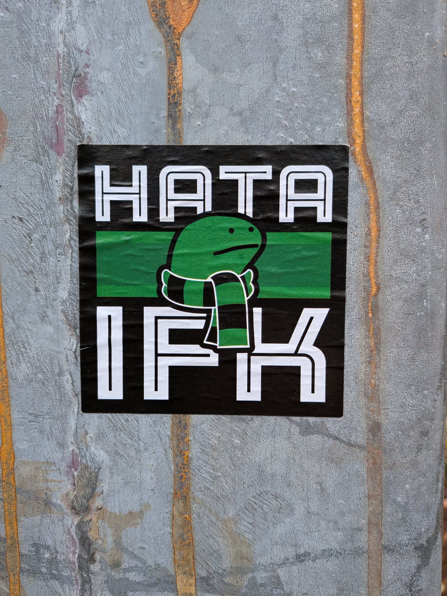Street sticker The sticker is predominantly black with white text and a green and black striped scarf around a cartoonish green character. The text 'HATA' is at the top in large white block letters, and 'IFK' is at the bottom, also in large white block letters. Between the text, there is a horizontal green band. The character is green with small black dots for eyes and a simple mouth, wearing a scarf with alternating black and green stripes. The sticker appears to be slightly creased, indicating it is a physical sticker applied to a surface.