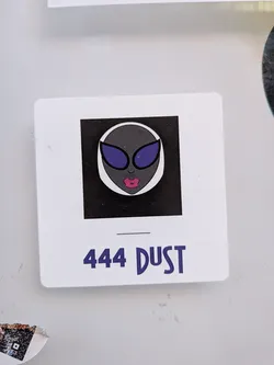 Street sticker The sticker is a white rectangle with rounded corners, resembling a slide or photo frame. It features a central black square containing a stylized alien head. The alien has dark grey skin, large, reflective purple eyes or glasses, and prominent, full pink lips. The alien's head is outlined in white against the black background. Below the central image, on the white border of the sticker, is the text '444 DUST' in a bold, purple, slightly rounded font, with a thin black horizontal line above it.