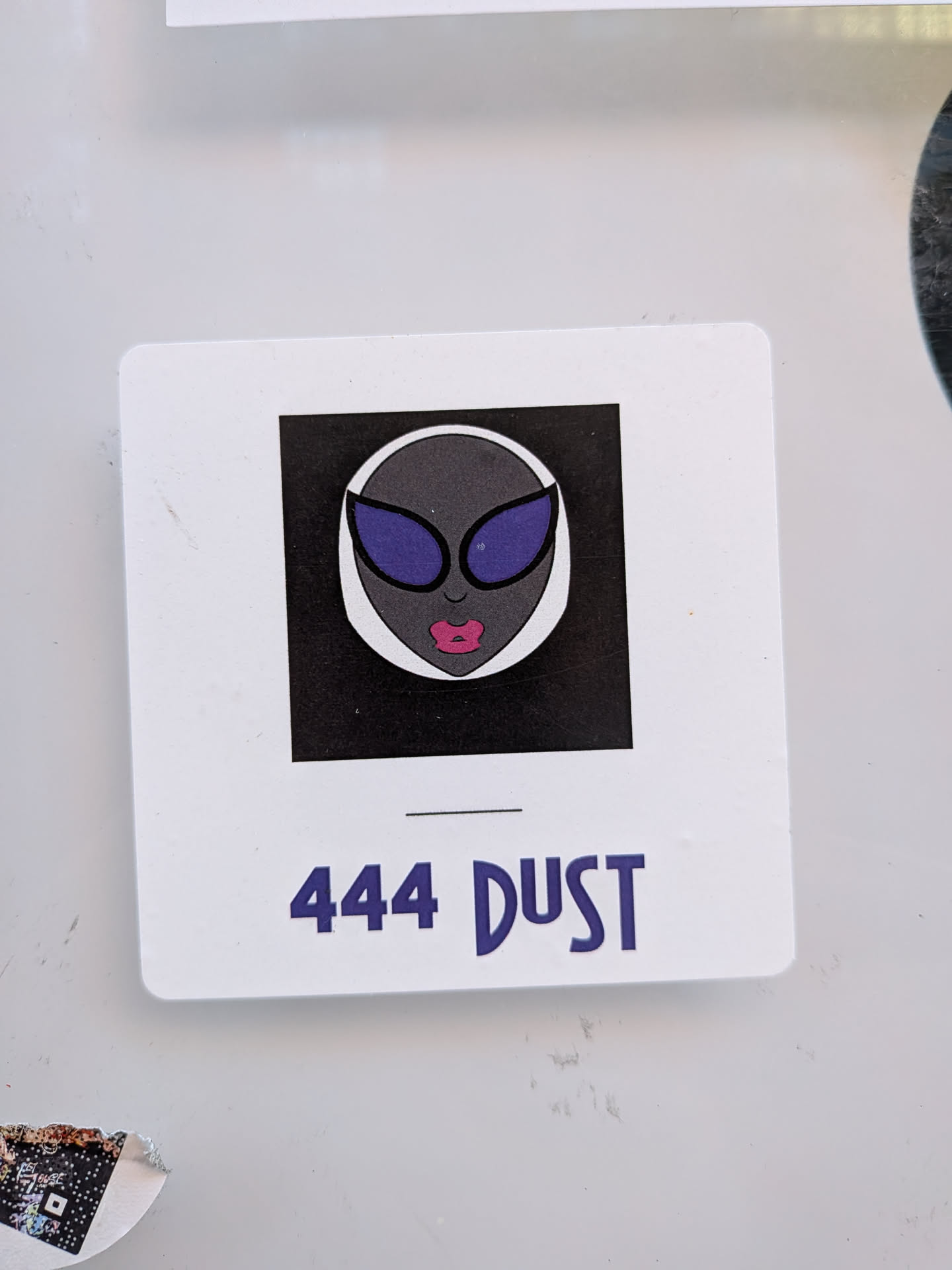 The sticker is a white rectangle with rounded corners, resembling a slide or photo frame. It features a central black square containing a stylized alien head. The alien has dark grey skin, large, reflective purple eyes or glasses, and prominent, full pink lips. The alien's head is outlined in white against the black background. Below the central image, on the white border of the sticker, is the text '444 DUST' in a bold, purple, slightly rounded font, with a thin black horizontal line above it.