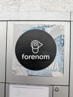 Street sticker The sticker is a circular, black decal showcasing a white outlined logo that appears to be a stylized combination of an eye and a location pin icon. Below the logo, the word "forenom" is printed in white lowercase letters. The sticker shows signs of wear, with some peeling and remnants of an older blue and white sticker visible around its edges.