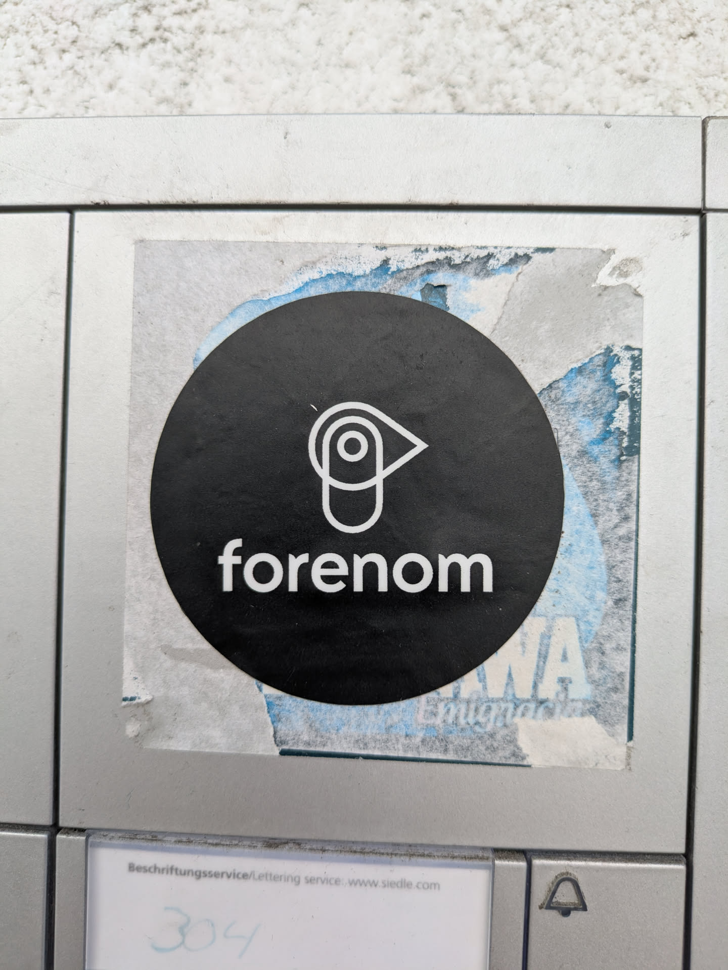 The sticker is a circular, black decal showcasing a white outlined logo that appears to be a stylized combination of an eye and a location pin icon. Below the logo, the word "forenom" is printed in white lowercase letters. The sticker shows signs of wear, with some peeling and remnants of an older blue and white sticker visible around its edges.