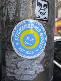 Street sticker The sticker is round with a light blue background and yellow concentric circles. The text 'STICKERBOMB WORLD' is written in a circle around the edge. Inside the yellow circles is a cartoon bomb with 'X's for eyes and its tongue sticking out. The text 'EST 2008' is below the bomb. Additional website and social media information is written around the outer edge of the sticker.