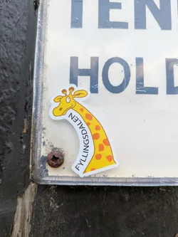 Street sticker A vibrant yellow cartoon giraffe sticker with orange spots and brown outlines is prominently displayed on a worn, white sign. The giraffe's head, featuring two small ossicones and a friendly expression, is at the top, and its long, curving neck extends downwards. Along the curve of its neck, the black text 'FYLLINGSDALEN' is written vertically. The sticker has a clean white border. The background sign shows faint, large black letters, with 'EN' and 'HOLD' being partially visible. A rusty screw is seen below the giraffe's neck, securing the sign. The overall setting appears to be an outdoor urban environment, with a dark, textured surface (possibly a wall or post) on the left.