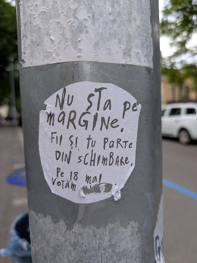 A sticker with the text 'Nu sta pe margine! Fii și tu parte din schimbare. Pe 18 mai votăm' (Don't stay on the sidelines! Be part of the change. We vote on May 18th) affixed to a metal pole.