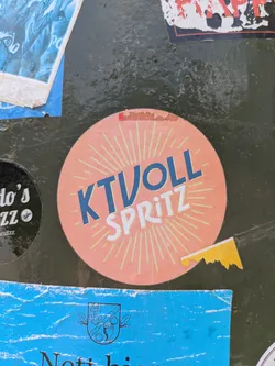 Street sticker The central sticker is circular with a peach-orange background. It features a bright, light yellow sunburst or starburst pattern emanating from the center. Overlaid on this pattern, in bold blue capital letters, is the word "KTVOLL", and directly below it, in smaller white capital letters, is "SPRITZ". The sticker shows some signs of wear and tear, especially around the edges, and a small, jagged yellow and white piece of another sticker is partially adhered to its bottom right edge. The sticker is placed on a dark, weathered, and textured green surface, possibly metal, alongside other partial stickers.
