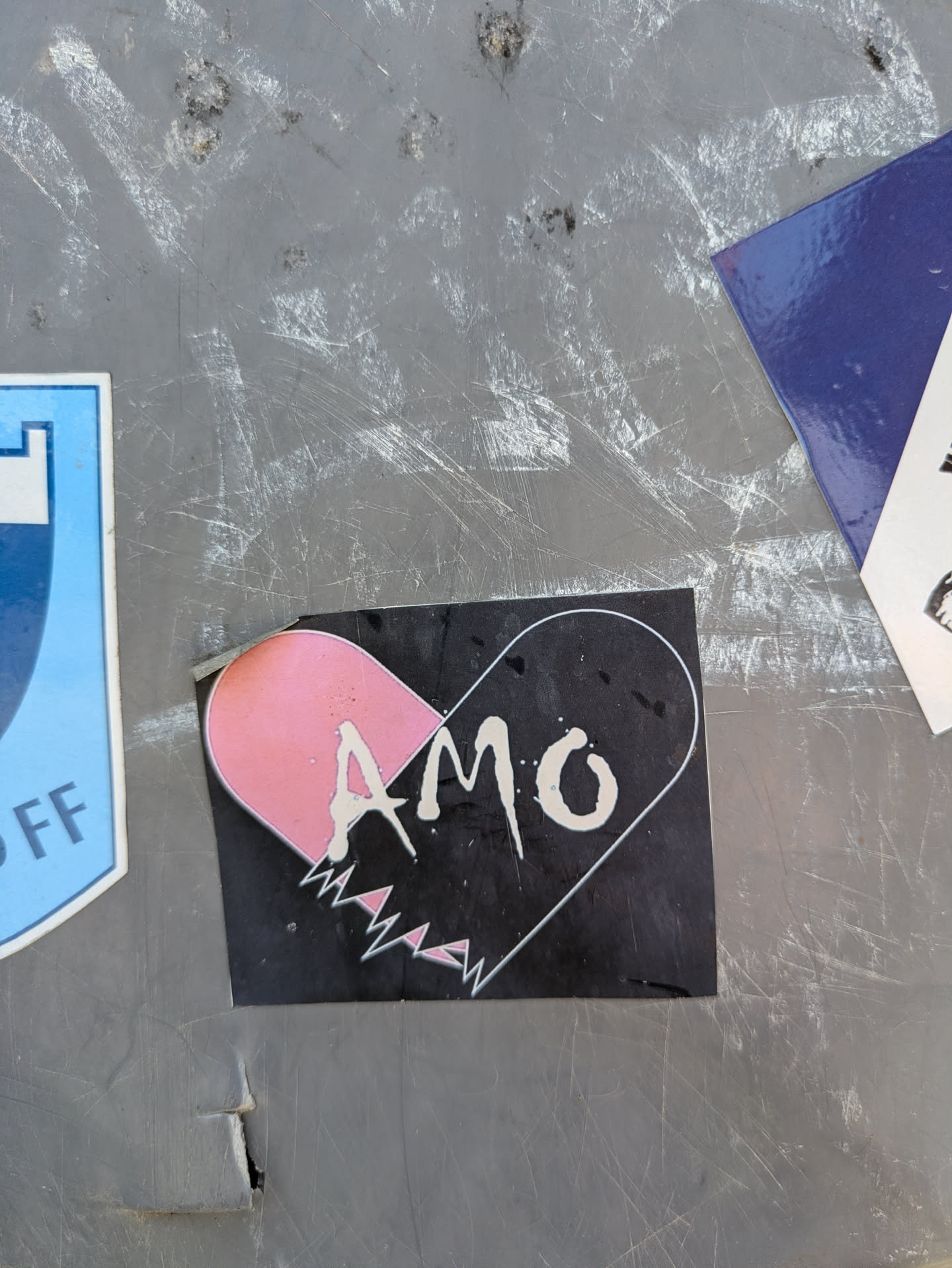 A black sticker featuring a stylized broken heart design. The upper left portion of the heart is filled with a light pink color, while the rest of the heart is outlined in white on a black background. The word "AMO" is written in white, stylized letters across the center of the heart. The bottom of the heart is jagged, resembling a broken edge.