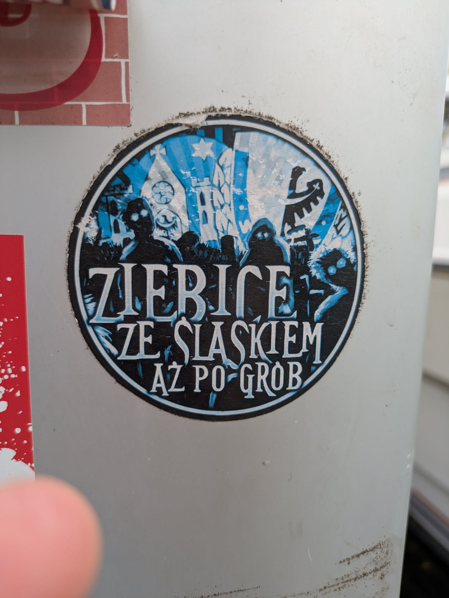 This is a circular sticker, prominently displaying dark, silhouetted figures against a textured blue and white background. The central text, 'ZIERICE ZE SLASKIEM AZ PO GROB,' is rendered in white uppercase letters, occupying a significant portion of the sticker. The imagery includes a zombie-like figure with glowing eyes, a hooded figure, and another creature with glowing eyes. In the background, elements resembling a gothic church or cathedral, a flagpole with a banner (possibly featuring an eagle), and other architectural details are visible. The sticker has a worn appearance, with some peeling and dirt around its edges, suggesting it has been exposed to the elements.