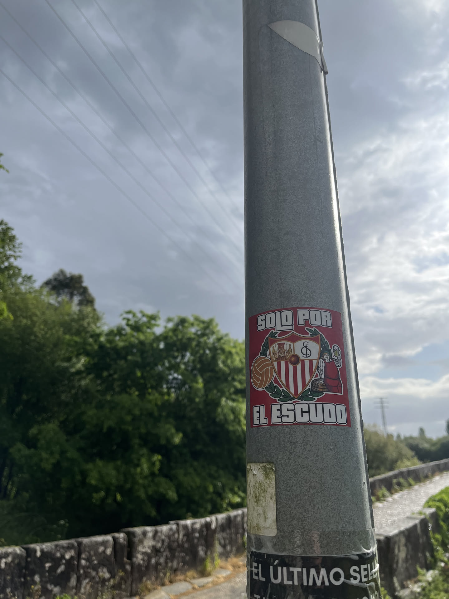 A rectangular sticker with a prominent red background, affixed to a grey metal pole. The central feature is a shield-shaped crest, primarily in red and white. The lower half of the crest displays vertical red and white stripes. The upper half is divided into three sections: on the left, three figures (possibly saints or historical figures); in the center, a vintage-style football with a crown above it and the intertwined letters 'SFC'; and on the right, a figure resembling King Ferdinand III of Castile. The crest is flanked by green laurel-like branches. To the left of the crest, there's a separate depiction of a classic brown football. To the right, a cartoonish figure, possibly a woman in traditional attire, is shown holding a large, frothy beer mug. Above the crest, in white block letters, is the text 'SOLO POR', and below the crest, also in white block letters, is 'EL ESCUDO'. The sticker shows some signs of wear and minor dirt accumulation, particularly along its edges.