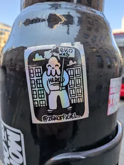 Street sticker A rectangular sticker with a black background and a holographic, iridescent finish for its main elements. It features a stylized, blob-like character with a round head, two small eyes, and three wavy lines resembling tentacles for a mouth. The character has a simple body and legs, and is holding a stick or pole in its right hand. Above its head, it holds a sign that reads 'EKO WAS', and on its chest, the word 'HERE' is written. Behind the character are two minimalist, outlined representations of multi-story buildings. The entire design is outlined in black, and the holographic material reflects various pastel colors like blue, green, pink, and yellow. At the bottom center of the sticker, a social media handle is visible. The sticker shows some signs of wear and slight peeling around its edges.