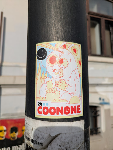 A sticker depicting a surreal cartoon bunny with wide eyes and a slightly manic expression, munching on what appears to be bananas. The sticker is on a pole. There's a smiley face in the corner and a tag at the bottom.