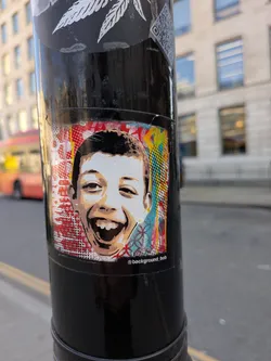 Street sticker The central sticker features a stylized, high-contrast portrait of a young child's face, captured mid-laugh with a wide-open mouth and crinkled eyes. The face itself is rendered in shades of white, light beige, and dark brown for outlines and shadows, giving it a stencil-like appearance. The background is a dynamic and colorful abstract composition, primarily featuring a red area with a white halftone dot pattern on the left, transitioning into vibrant splashes of yellow, blue, and green on the right. The blue/green section includes a subtle geometric or floral pattern. The overall aesthetic is energetic and joyful, reminiscent of pop art or street art.