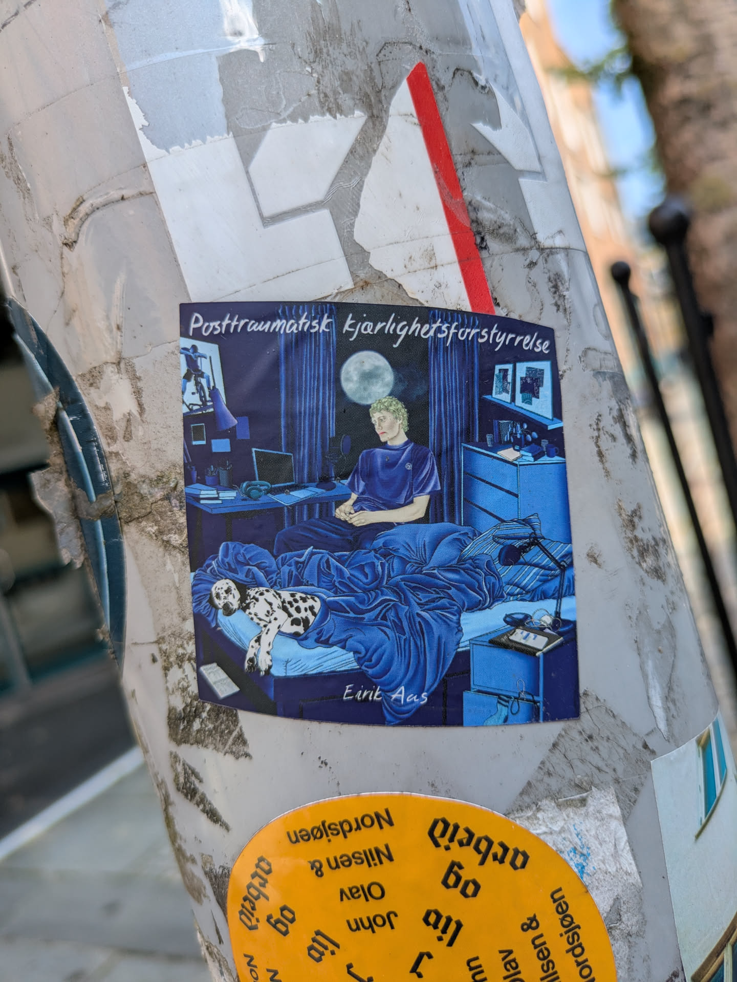 The sticker features a detailed illustration predominantly in shades of blue. It depicts a person with light, possibly curly hair, sitting upright in a bed with blue blankets. They appear pensive, looking towards the left side of the frame. A Dalmatian dog is curled up and sleeping under the blankets beside the person. The background shows a bedroom or studio setting, complete with a desk holding a laptop, headphones, and a microphone, suggesting a creative or online activity. A dresser and framed pictures adorn the wall, and a large full moon is visible through a window, casting light into the room. A lamp sits on the bedside table. The overall atmosphere is quiet and contemplative.