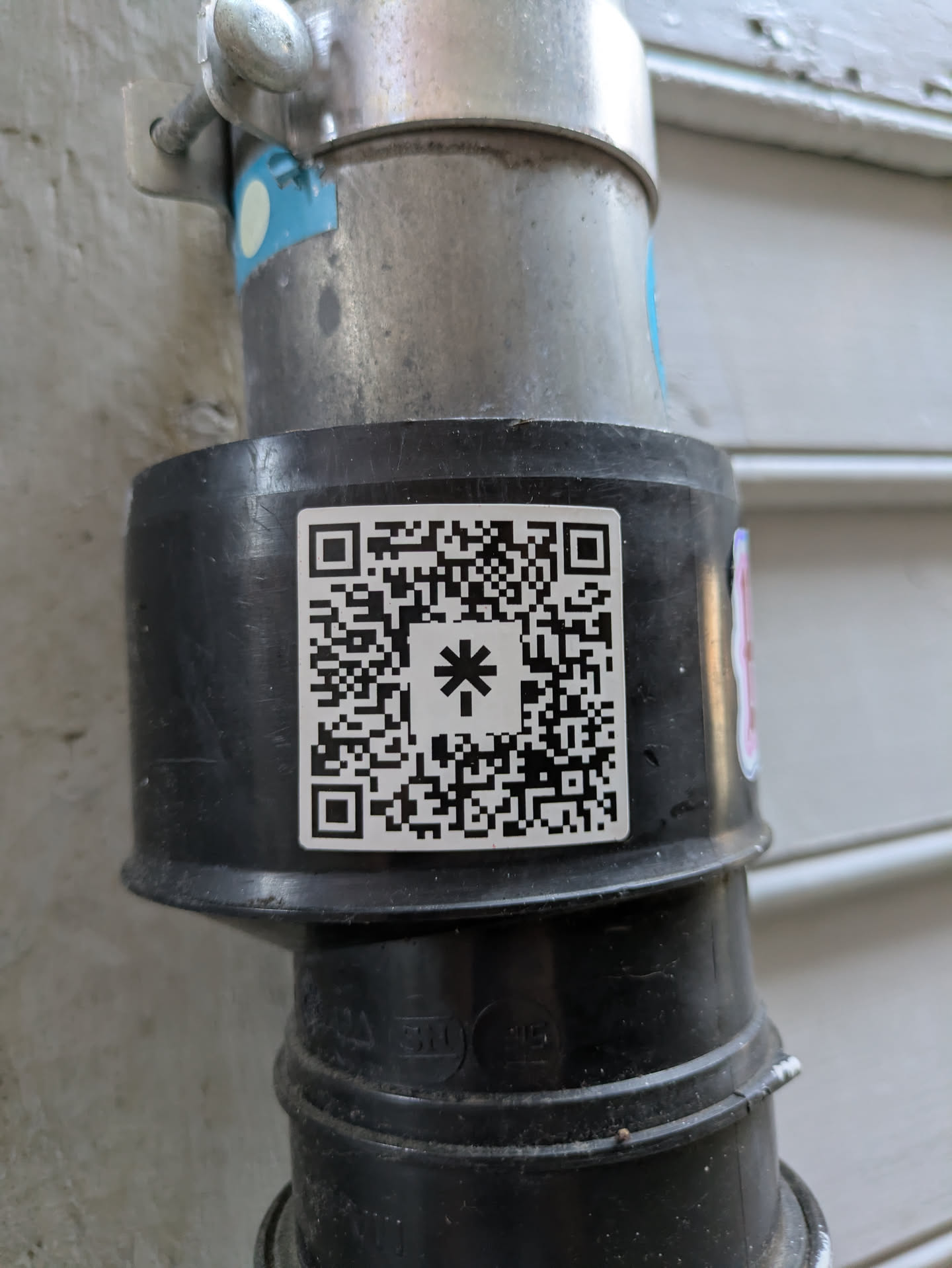 A white square sticker featuring a black QR code is prominently displayed on a dull black pipe, which is part of a larger plumbing or conduit system. The QR code contains a large, stylized asterisk (*) symbol in its center, adding a unique visual element to the standard code pattern. The pipe is connected to a larger metallic pipe assembly above it, which is secured by a metal clamp. The background shows a light-colored building surface, possibly stucco or siding.