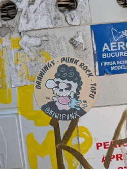 Street sticker A beige circular sticker featuring a cartoon skull with curly black hair, bulging bloodshot eyes, and a pink tongue spitting blue liquid. Text arches around the top border reading 'DRAWINGS - PUNK ROCK - TOFU', with the handle '@KIWIPUNX' on the character's shirt.