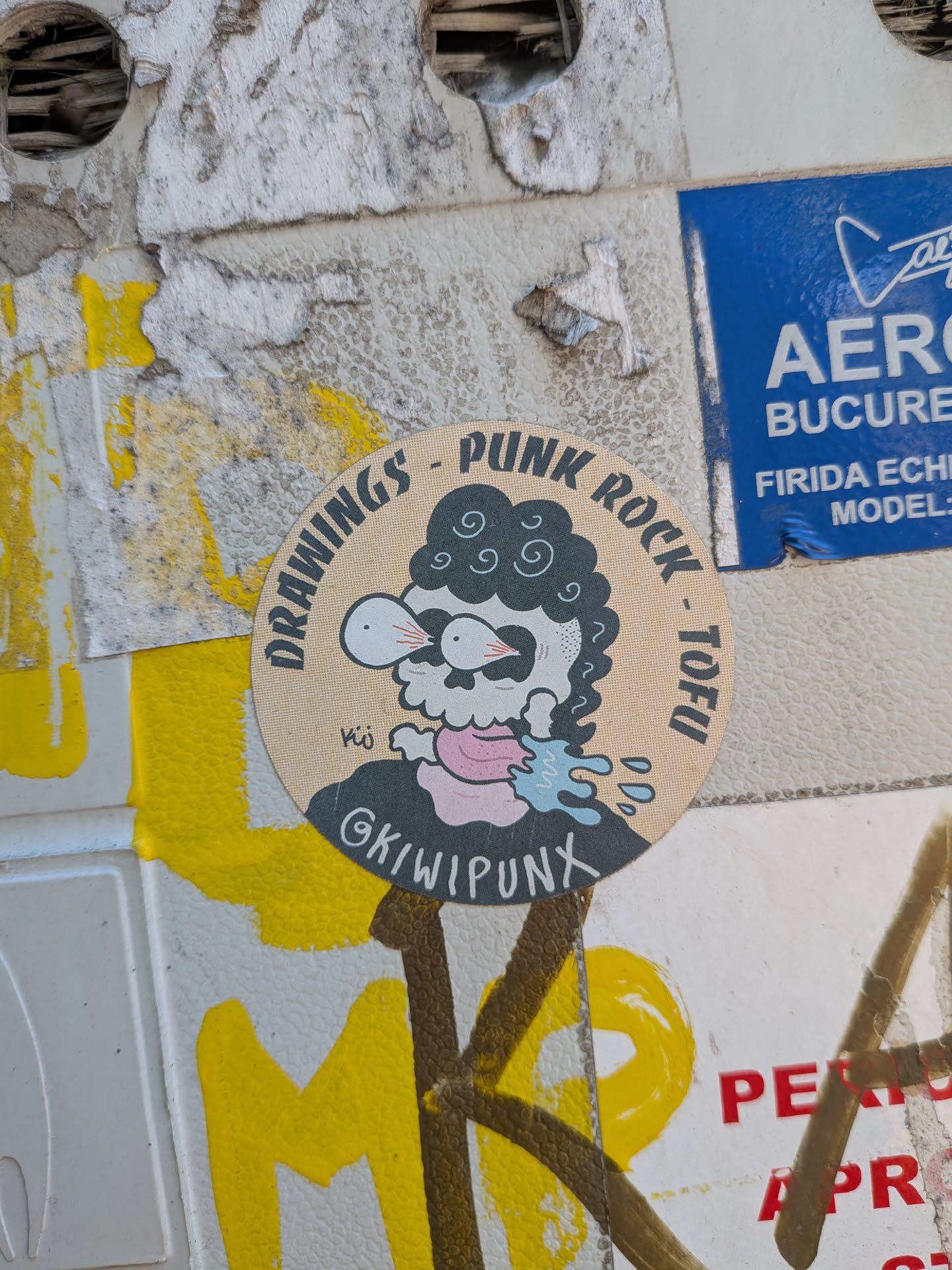 A beige circular sticker featuring a cartoon skull with curly black hair, bulging bloodshot eyes, and a pink tongue spitting blue liquid. Text arches around the top border reading 'DRAWINGS - PUNK ROCK - TOFU', with the handle '@KIWIPUNX' on the character's shirt.
