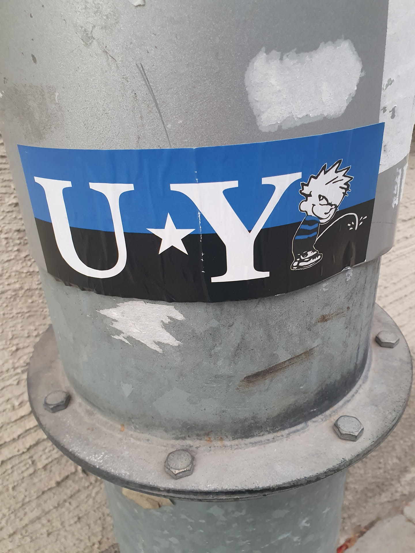 The sticker is horizontally oriented and features a blue, black, and white color scheme. The text 'U*Y' is prominently displayed in large white sans-serif font. To the right of the text is a cartoon character with spiky hair, wearing striped pants and sneakers, depicted in the act of urinating. The character has a mischievous expression. The sticker appears to be adhered to a metal pole.