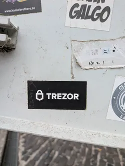 Street sticker A rectangular black sticker featuring a white stylized padlock icon next to the bold text 'TREZOR'.