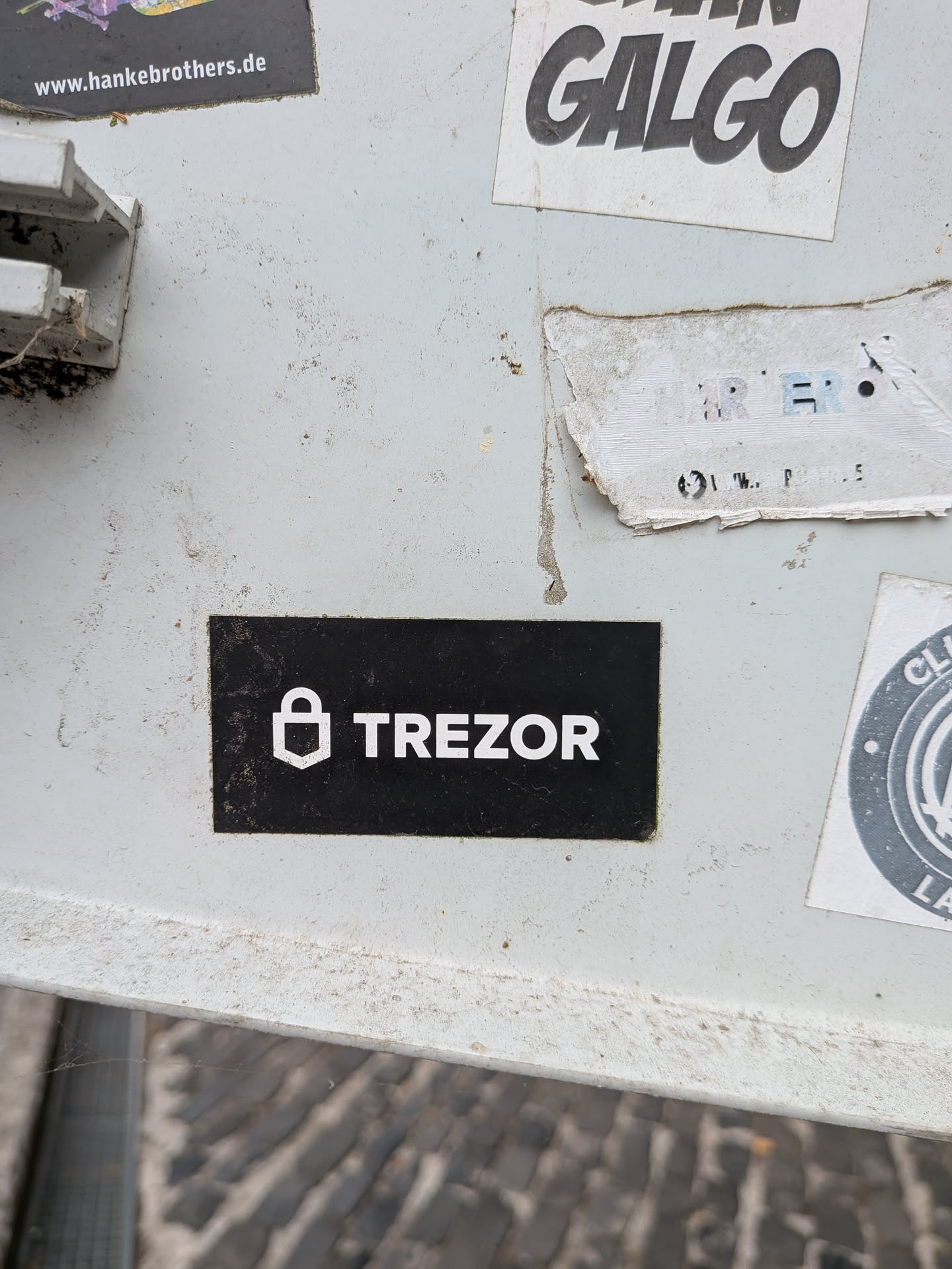 A rectangular black sticker featuring a white stylized padlock icon next to the bold text 'TREZOR'.