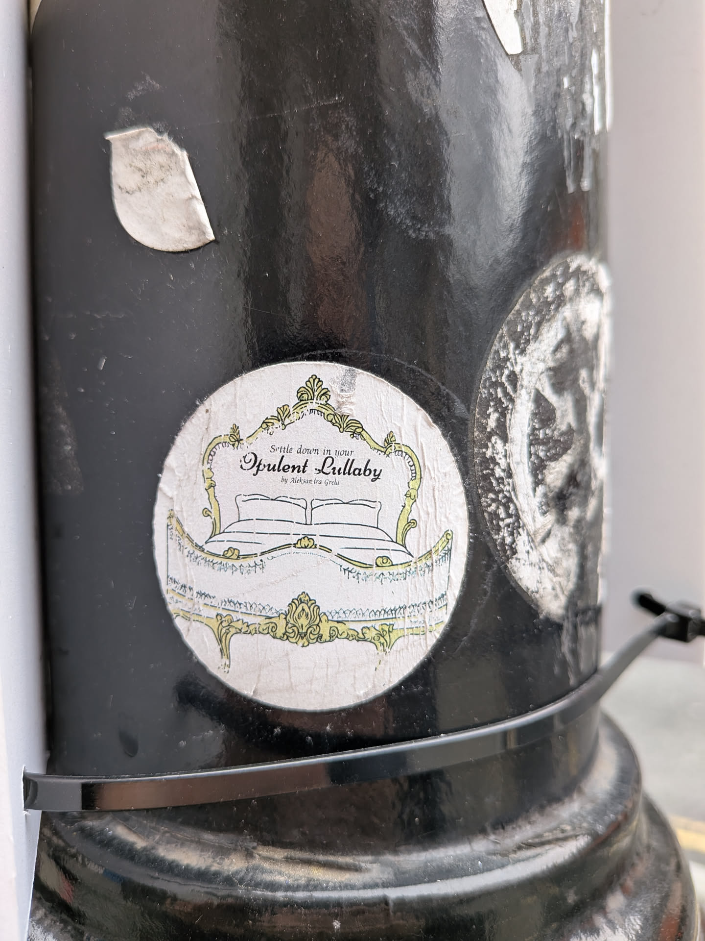 A circular sticker featuring an ornate, vintage-style bed frame with a headboard and footboard, rendered in a pale yellow or gold outline against an off-white, slightly textured background. The design has a baroque or rococo aesthetic with intricate scrolls and flourishes. Inside the bed frame, two pillows and a blanket are depicted with simple line art. Above the bed, the text 'Settle down in your Opulent Lullaby by Aleksandra Grelu' is written in a decorative script. The sticker appears slightly worn and creased, affixed to a black pole.