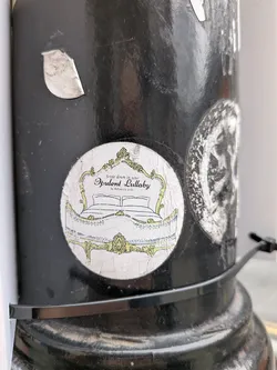 Street sticker A circular sticker featuring an ornate, vintage-style bed frame with a headboard and footboard, rendered in a pale yellow or gold outline against an off-white, slightly textured background. The design has a baroque or rococo aesthetic with intricate scrolls and flourishes. Inside the bed frame, two pillows and a blanket are depicted with simple line art. Above the bed, the text 'Settle down in your Opulent Lullaby by Aleksandra Grelu' is written in a decorative script. The sticker appears slightly worn and creased, affixed to a black pole.