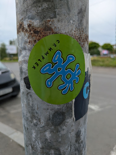 A round green sticker on a metal pole featuring a blue, abstract design in the center. The text "CRUMPLER" is written around the edge of the sticker. The pole shows signs of rust and wear.