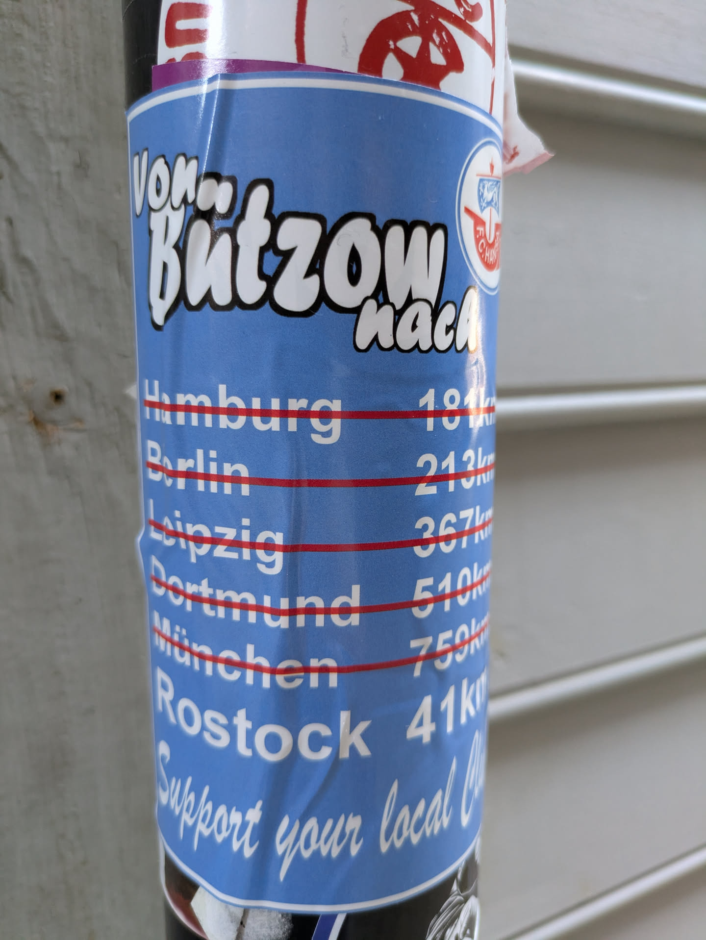 The sticker is vertically oriented, primarily blue with white text and a red line marking each city's distance. At the top, it reads 'Von Butzow nach'. Below this, a list of cities and their distances from Butzow is displayed: Hamburg 181km, Berlin 213km, Leipzig 367km, Dortmund 510km, München 759km, and Rostock 41km. The FC Hansa Rostock logo is visible in the top right section. At the bottom, a call to action reads 'Support your local Club'. The sticker is adhered to a pole.
