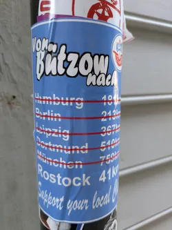 Street sticker The sticker is vertically oriented, primarily blue with white text and a red line marking each city's distance. At the top, it reads 'Von Butzow nach'. Below this, a list of cities and their distances from Butzow is displayed: Hamburg 181km, Berlin 213km, Leipzig 367km, Dortmund 510km, München 759km, and Rostock 41km. The FC Hansa Rostock logo is visible in the top right section. At the bottom, a call to action reads 'Support your local Club'. The sticker is adhered to a pole.