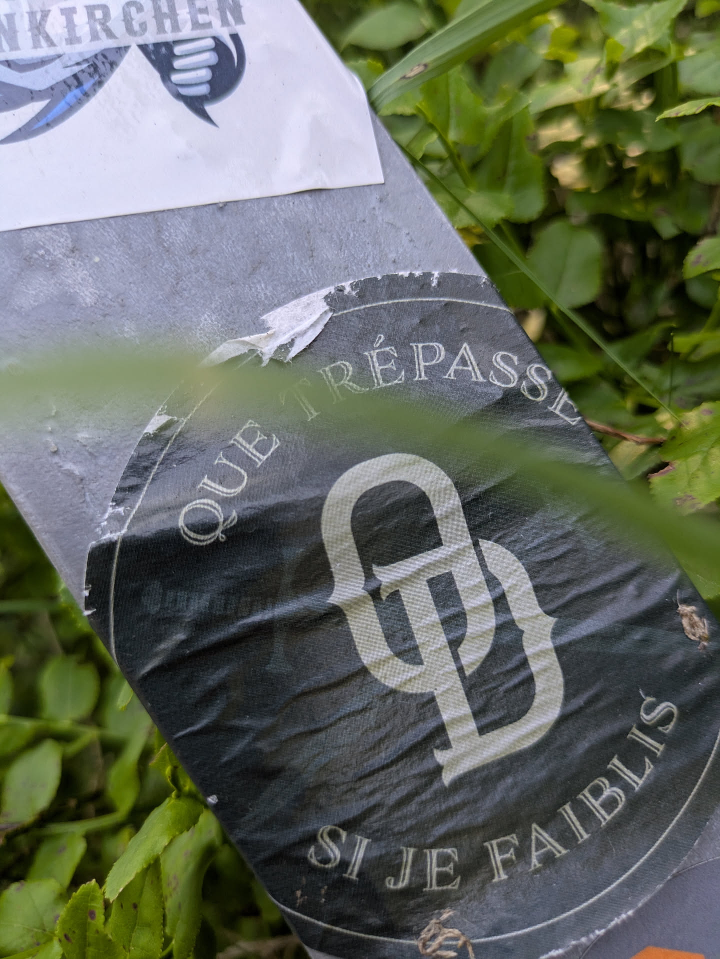 A dark, circular sticker affixed to a gray surface, partially obscured by green foliage. The sticker features white, stylized text that reads 'QUE TRÉPASSE' along the top curve and 'SI JE FAIBLIS' along the bottom curve. In the center, there's a prominent, intricate white monogram, appearing to be intertwined letters, possibly 'GD' or similar in an old-style script. The sticker shows signs of wear and tear, with some edges torn.
