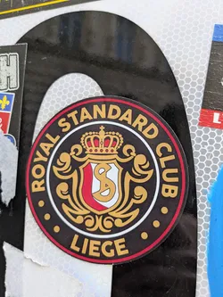 Street sticker This is a round sticker for the Royal Standard Club Liege, a football club. The sticker features the club's emblem, which includes a crown, a shield with the letters "S" and "C", and decorative flourishes. The text around the emblem reads "ROYAL STANDARD CLUB" at the top and "LIEGE" at the bottom.