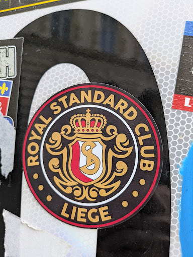 This is a round sticker for the Royal Standard Club Liege, a football club. The sticker features the club's emblem, which includes a crown, a shield with the letters "S" and "C", and decorative flourishes. The text around the emblem reads "ROYAL STANDARD CLUB" at the top and "LIEGE" at the bottom.