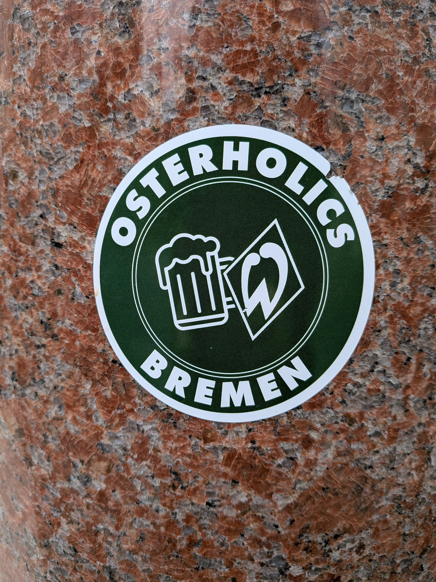 A circular sticker with a dark green background and a white border. The top arc of the circle features the word "OSTERHOLICS" in bold white capital letters, and the bottom arc reads "BREMEN" in similar styling. The center of the sticker contains two white outlined icons: a frothy beer mug on the left and a stylized 'W' enclosed within a diamond shape on the right, reminiscent of the Werder Bremen football club logo. The sticker is adhered to a reddish-brown granite-like surface.