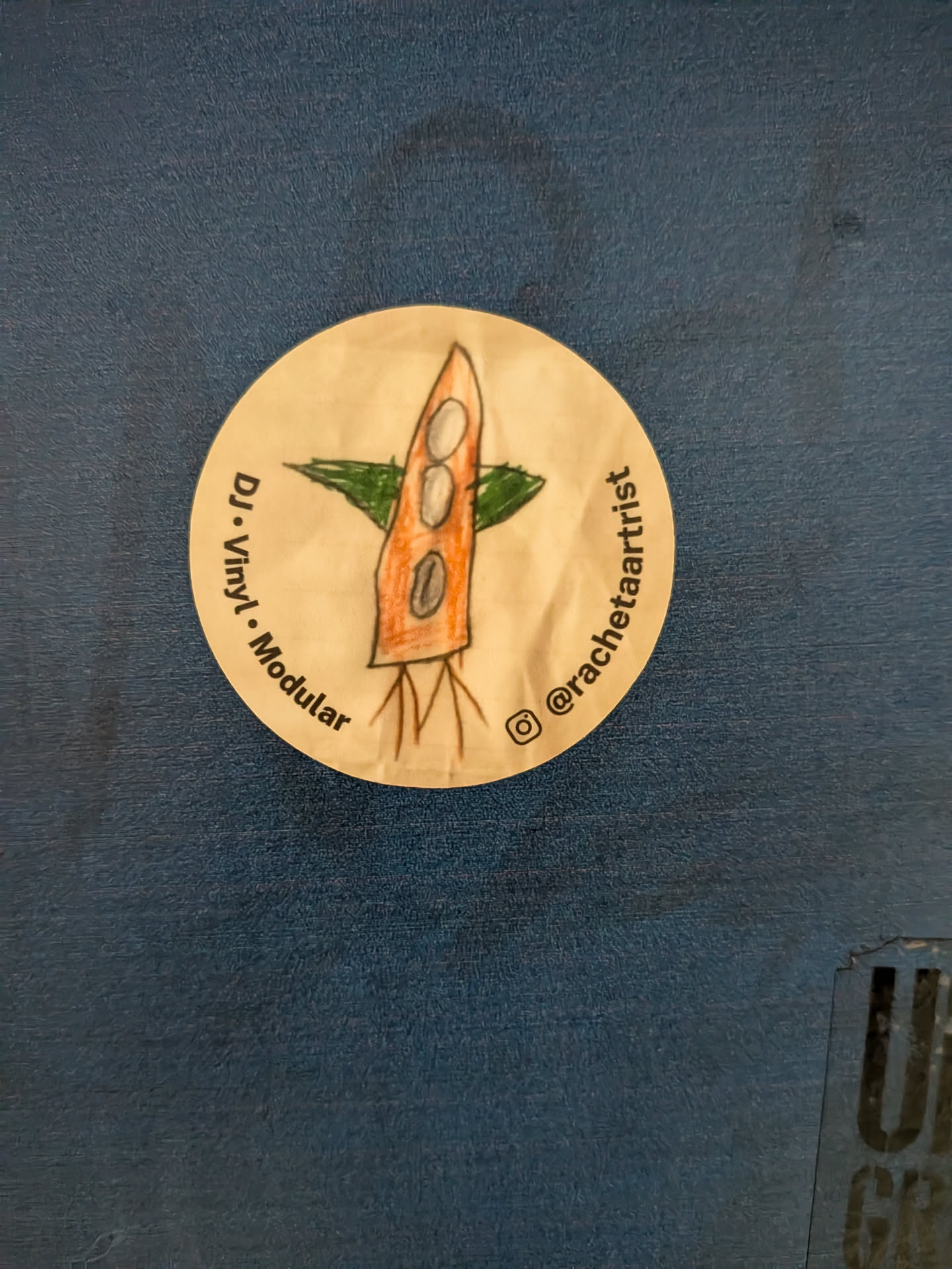 A round beige sticker displaying a hand-drawn orange rocket with green wings. Black text curves along the sides, including an Instagram handle.