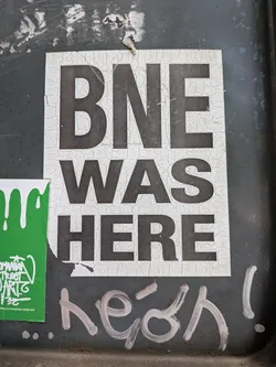 Street sticker A rectangular white sticker featuring bold black text 'BNE WAS HERE' stacked vertically. The sticker shows heavy cracking and weathering across its surface.