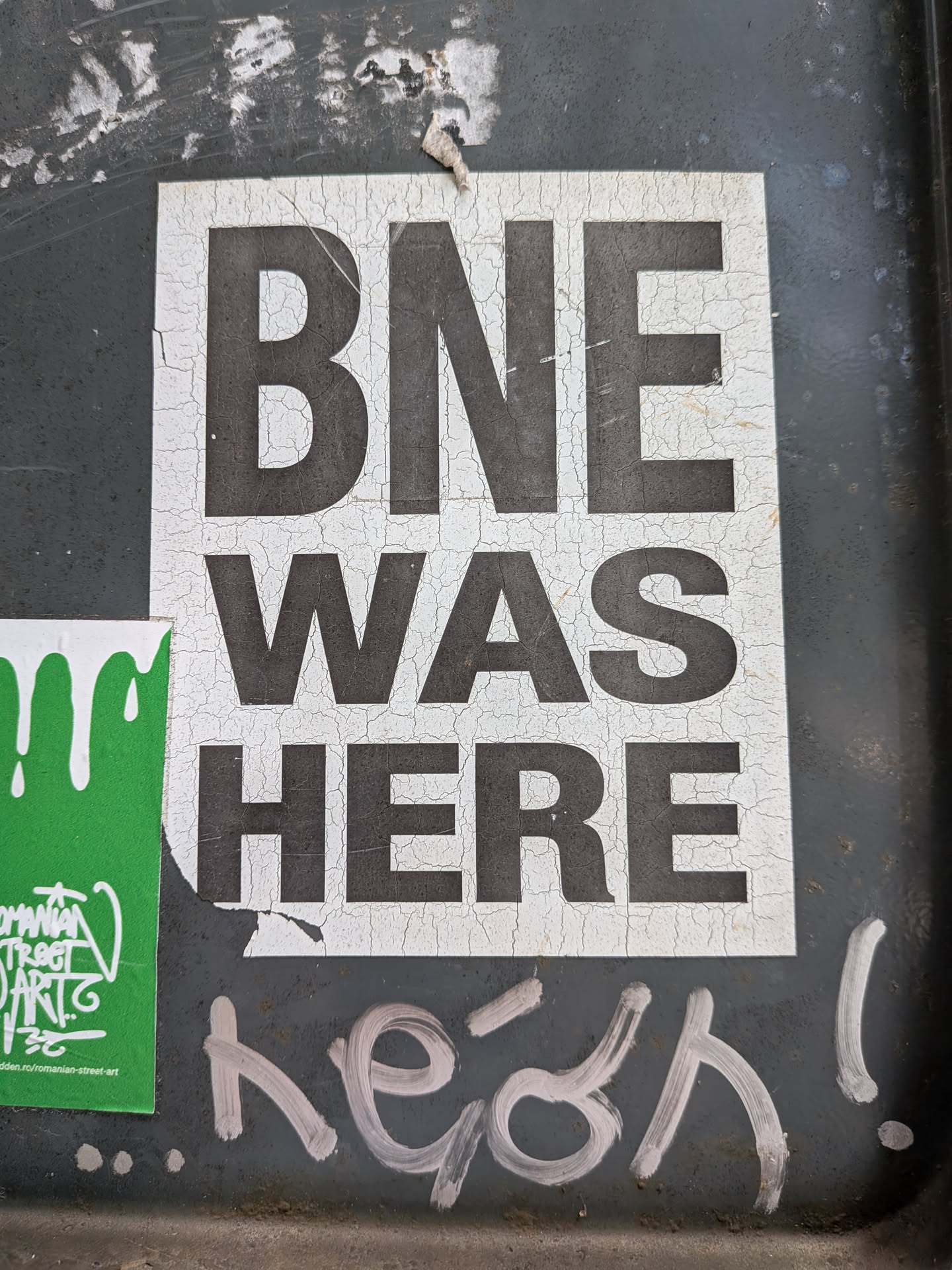 A rectangular white sticker featuring bold black text 'BNE WAS HERE' stacked vertically. The sticker shows heavy cracking and weathering across its surface.