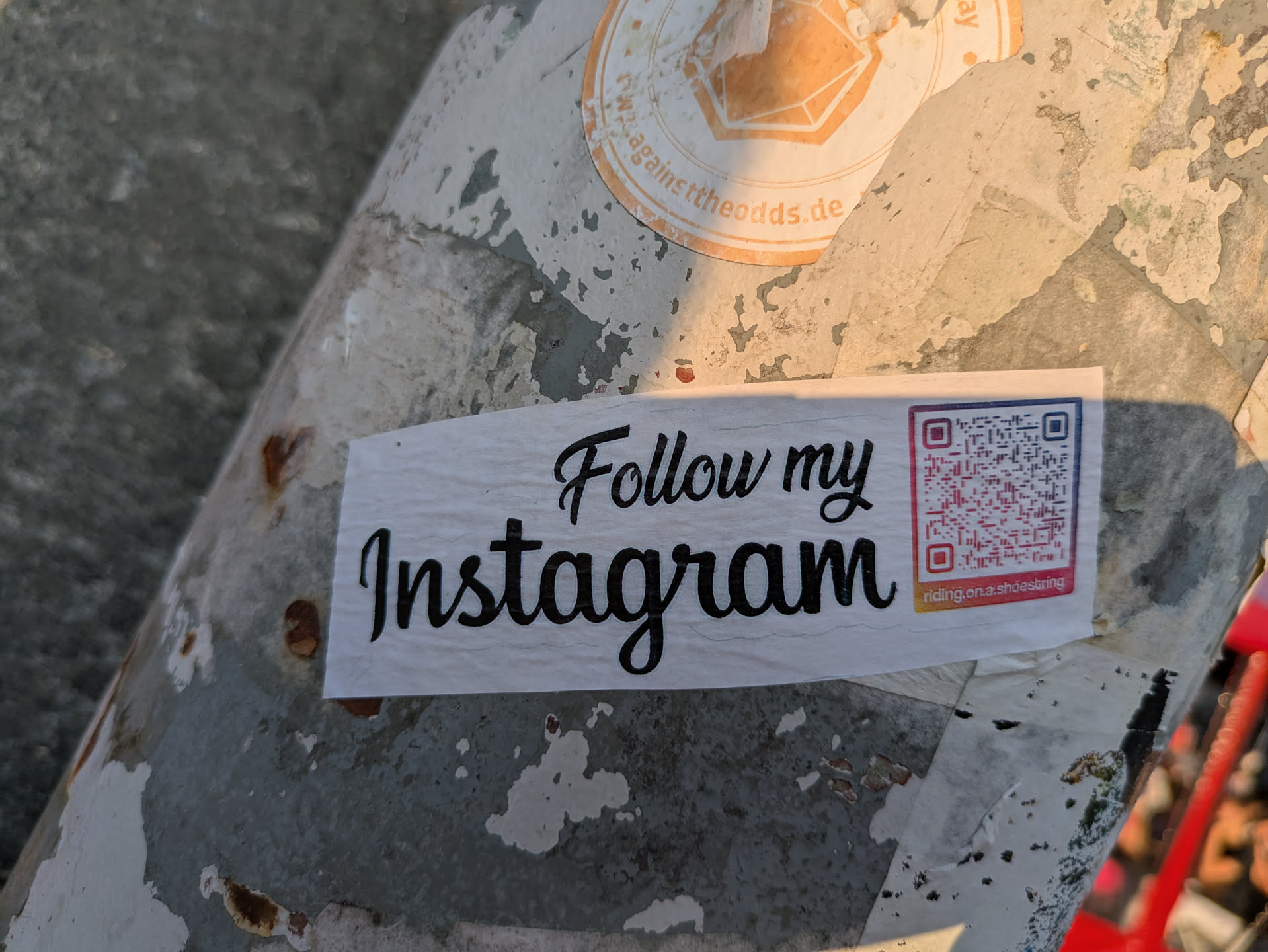 A rectangular white sticker with 'Follow my Instagram' in a large, elegant script font, placed on a weathered, peeling grey and white pole. To the right of the text, there is a colorful QR code. Below the QR code, the text 'riding.on.a.shoestring' is visible in a smaller font. The pole itself shows signs of rust and faded paint, with remnants of other old stickers partially visible in the background.