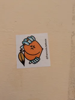 Street sticker An orange, fruit-like character sticker with a leaf is placed on a wall. The character has a gloomy expression with downturned eyebrows and mouth, as well as hands in the form of semicircles. A leaf is seen on the left side.
